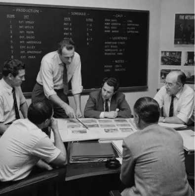 A vintage black and white photo of a film production crew in a meeting room reviewing printed materials on a table, with a large production schedule chalkboard visible in the background.