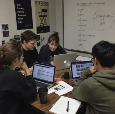 Four film students collaborating around laptops and notebooks in a classroom with a pre-production workflow whiteboard.