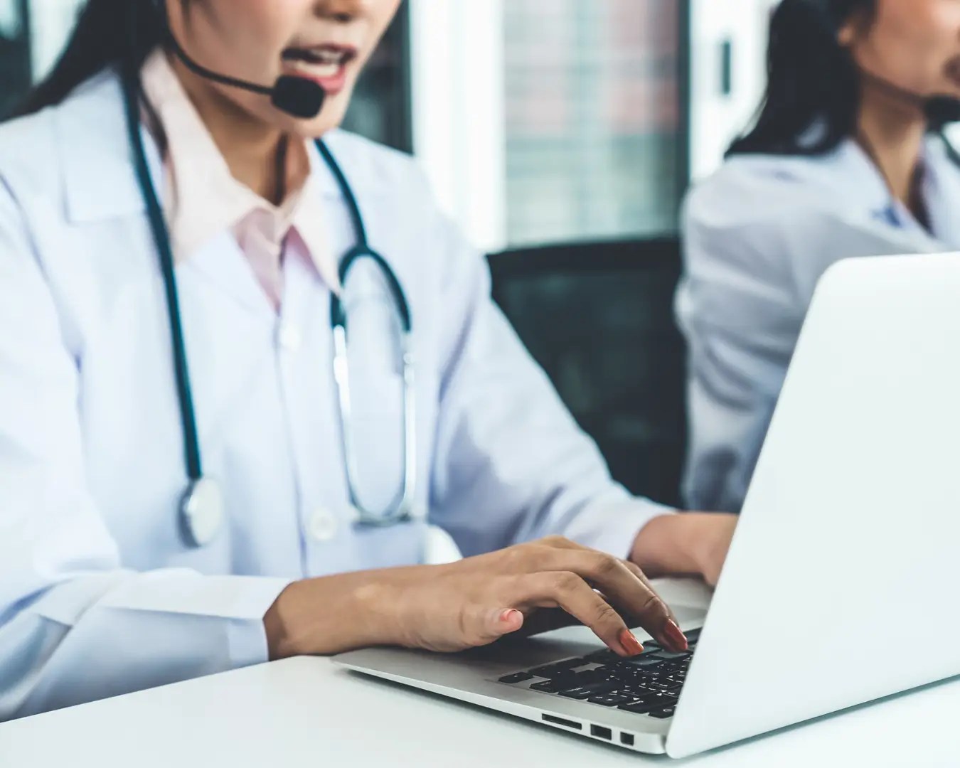 Woman typing on computer while talking on headset. She is wearing a white lab coat and stethoscope around her neck. Coworker can be seen sitting next to her.