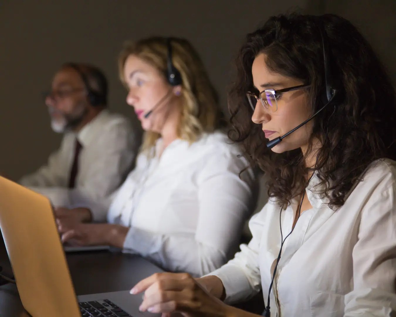 Two female adults and one male adult sitting at long desk with laptops in front of them, all wearing headsets.