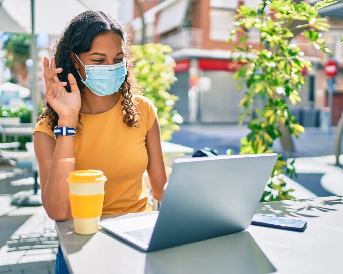 Young woman wearing face mask outside on a sunny summer day sitting at a coffee shop table in front of laptop, waving to screen.