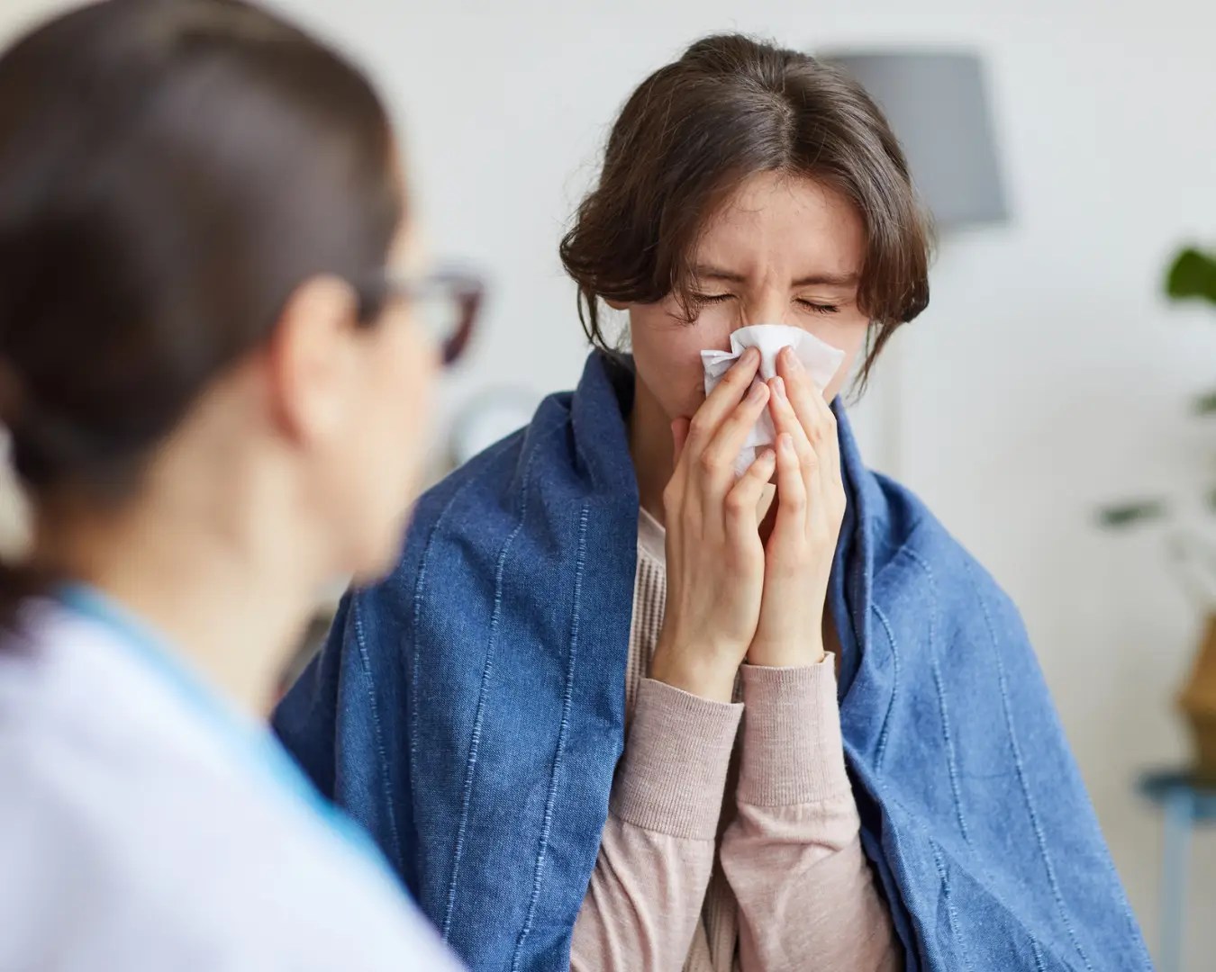 Young woman with blanket around her shoulders, blowing her nose, sitting across from female doctor.