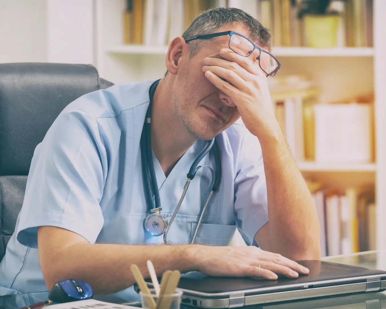 Exhausted male doctor sitting at his desk with his head in his hands.