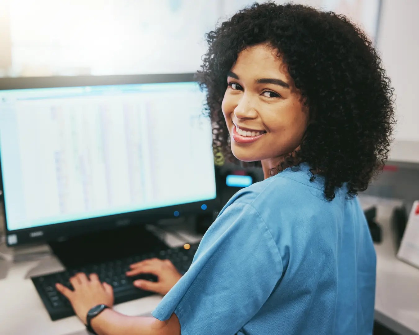 Young woman in scrubs sitting at desk working on computer, turned around smiling at camera.