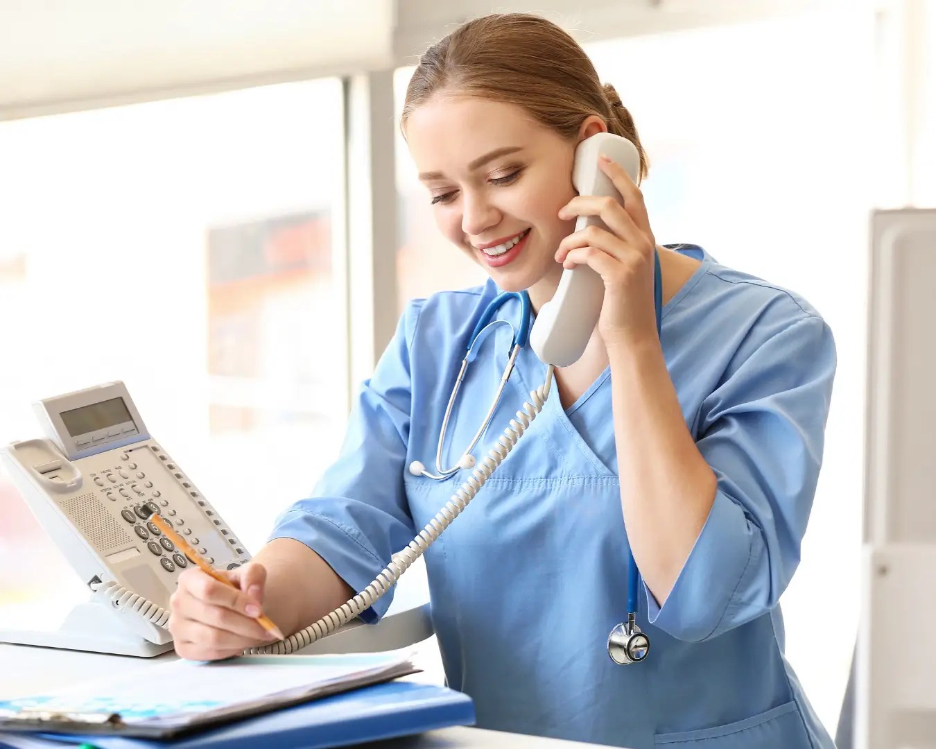 Young woman wearing scrubs, sitting at desk on the phone, writing in notepad.