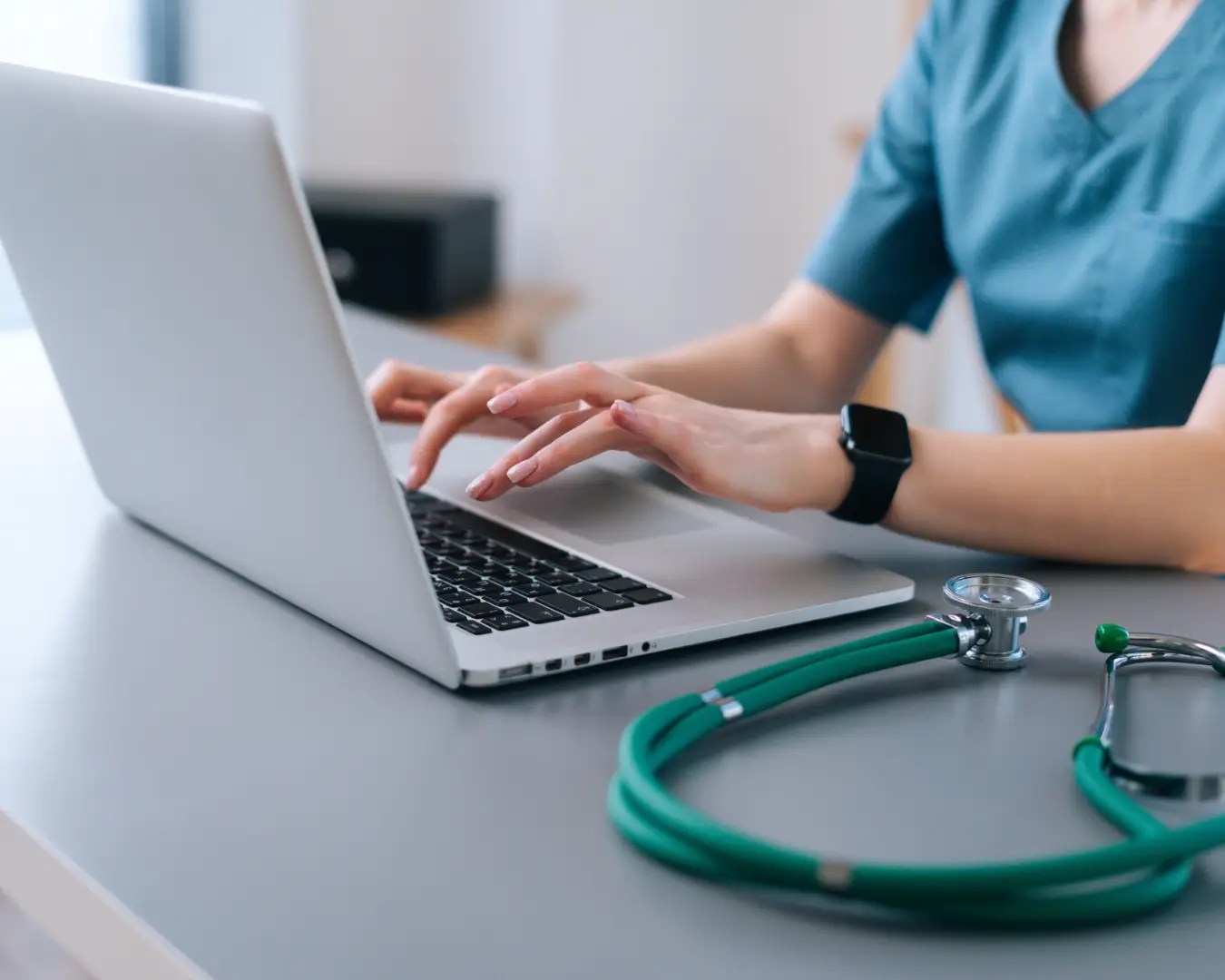 Close up of female doctor at desk typing on laptop. Stethoscope is resting next to laptop.
