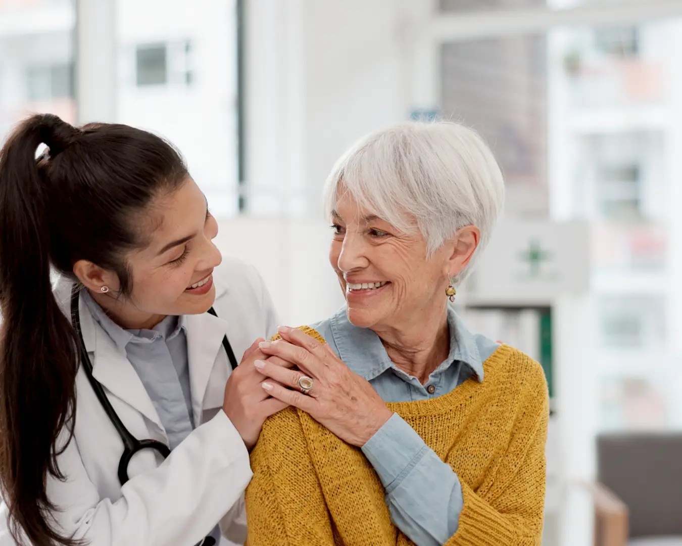 Mature woman in medical office with female doctor behind her with her hand on elderly woman's shoulder. Elderly woman has hand on top of female doctor's hand. Both smiling at one another.