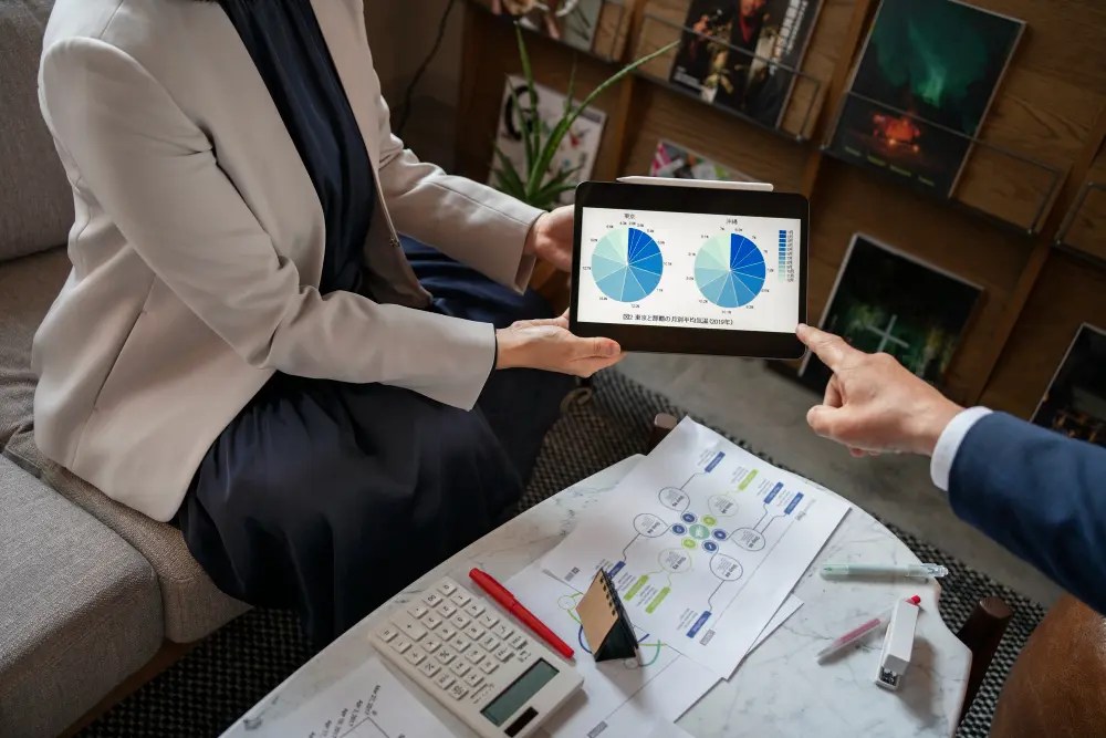 Two professionals are reviewing data analysis on a tablet while studying paper designs on a table.