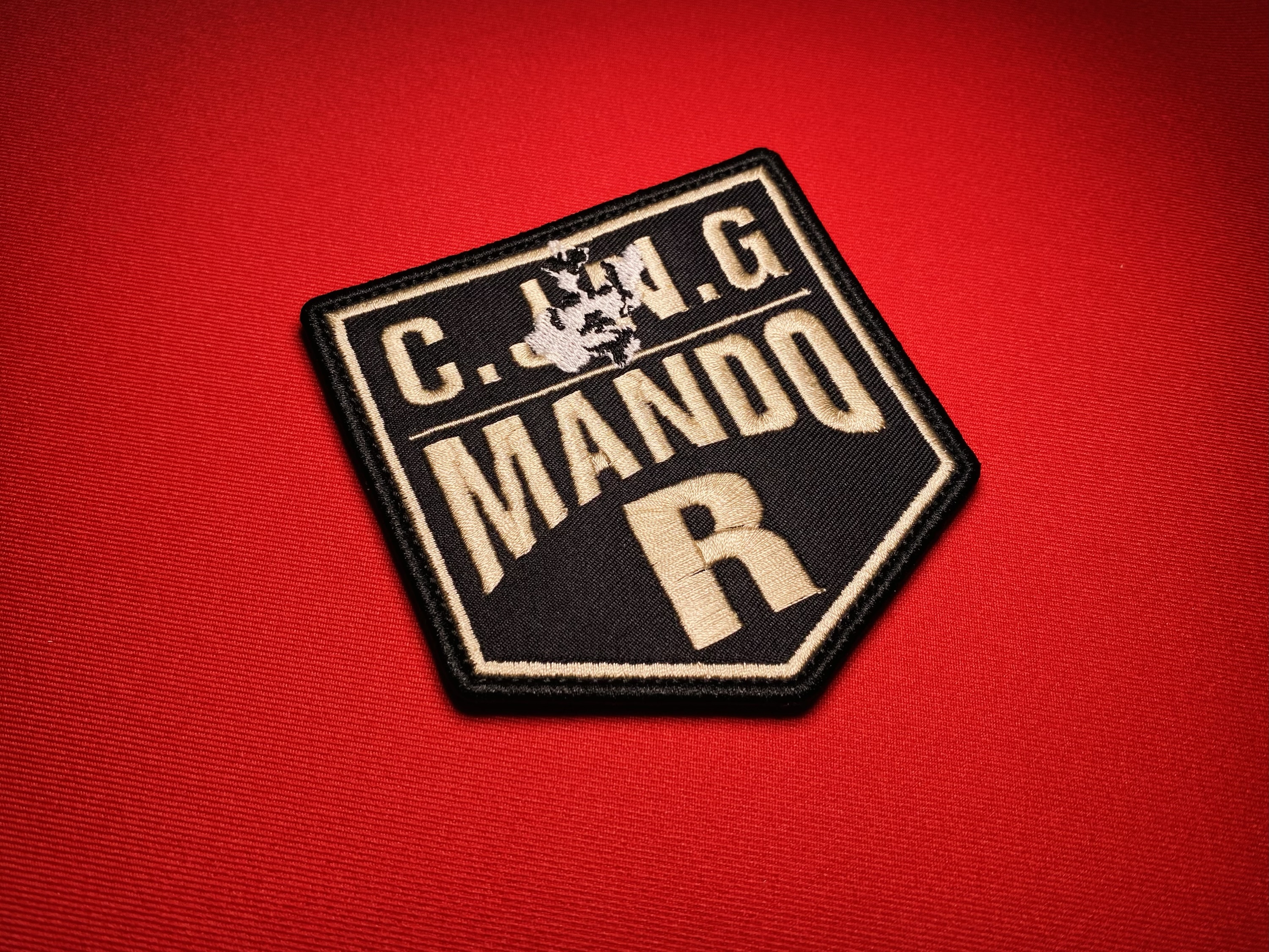 Embroidered black-and-tan shield patch reading “CJNG Mando R” with a Jalisco state outline above the text, photographed on a red fabric background.