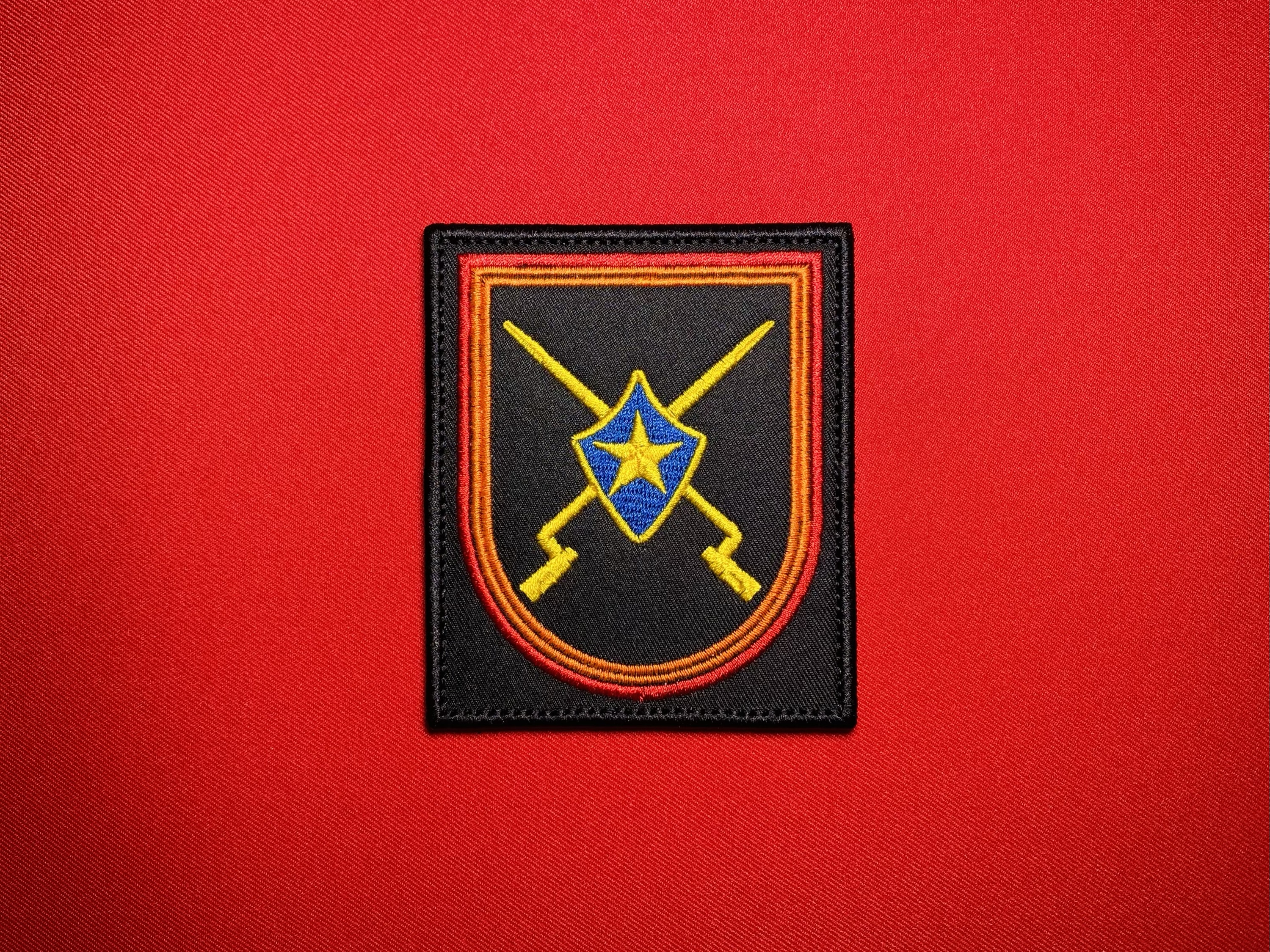 Front view of the full-color 114 OMSBr patch featuring a black field, red and orange border, blue shield, yellow star, and crossed weapons.