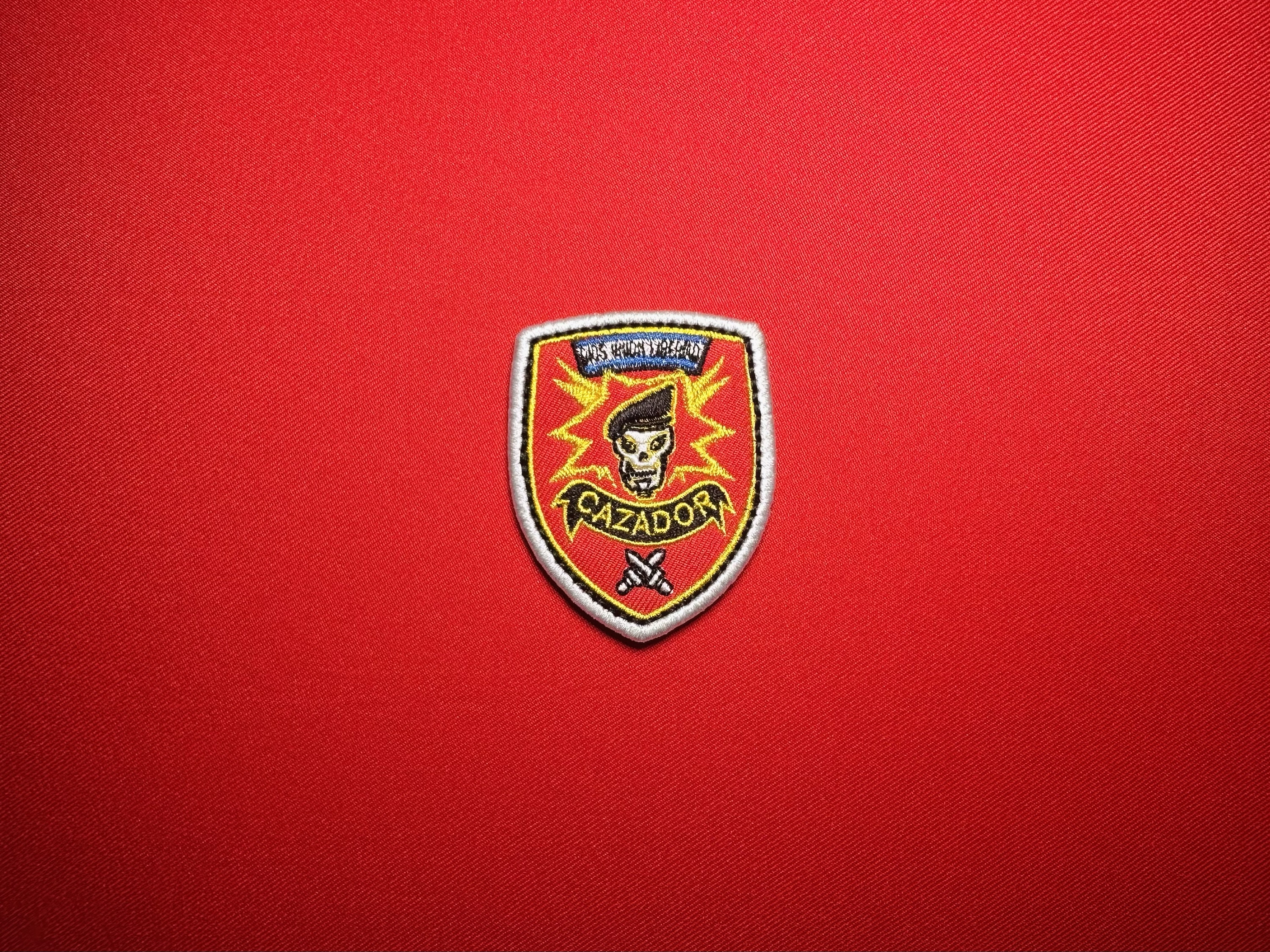 Salvadoran Cazador 2.5-inch embroidered shield patch featuring a MACV-SOG-style beret-wearing skull, yellow lightning bolts, red background, CAZADOR banner, and crossed weapons.