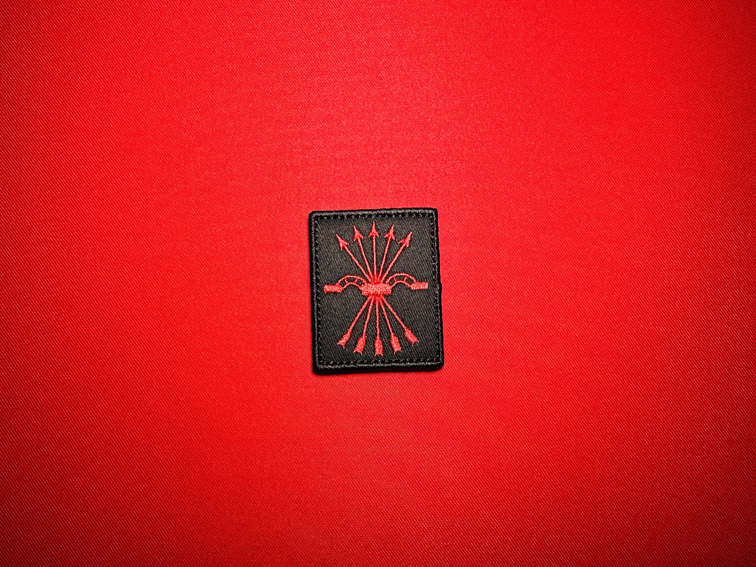 Black Falange Española replica patch featuring the red yoke and five arrows emblem, displayed on red fabric.
