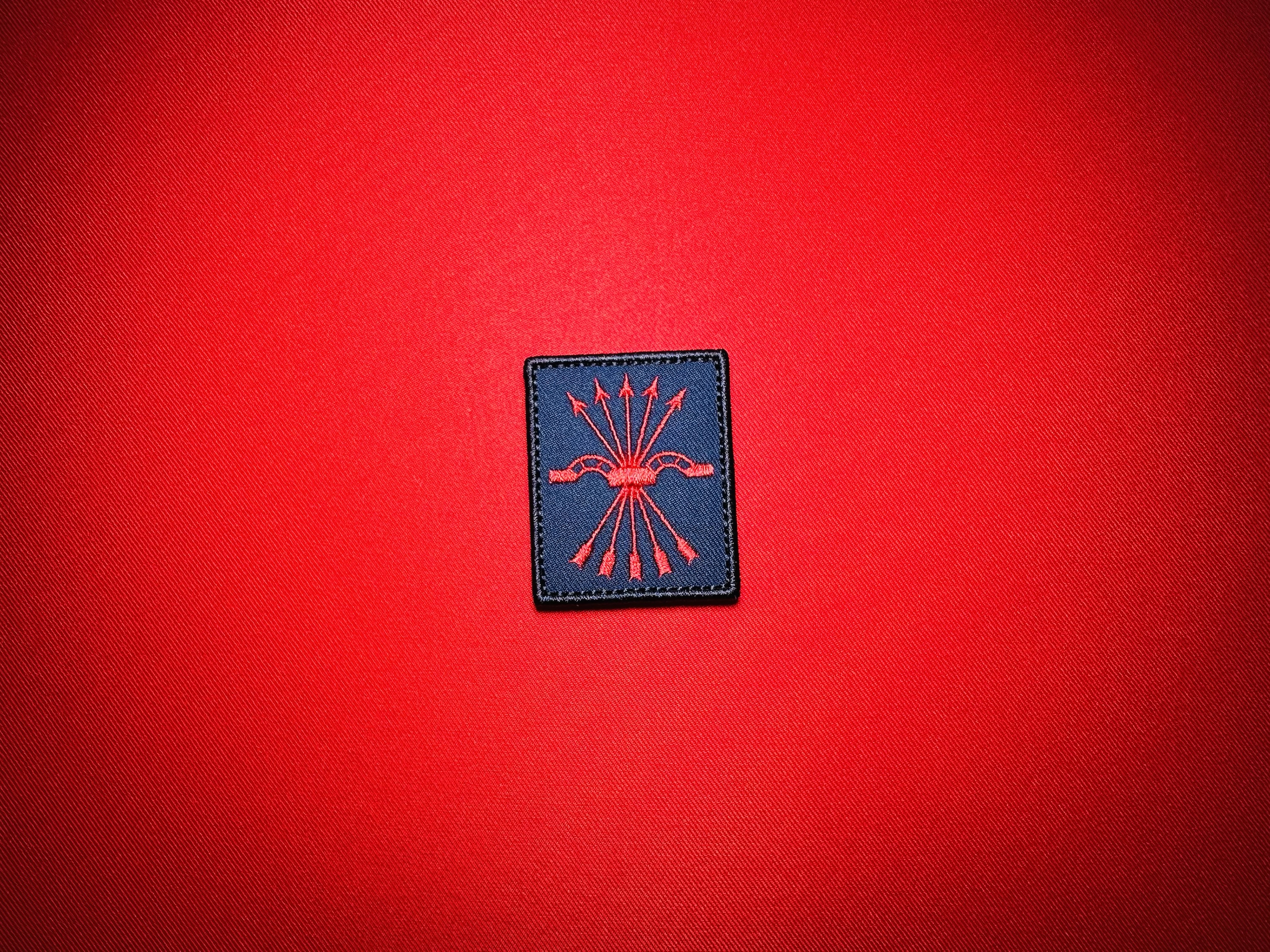 Spanish Falange replica patch with a red yoke and five arrows embroidered on dark blue cloth, displayed on red fabric.