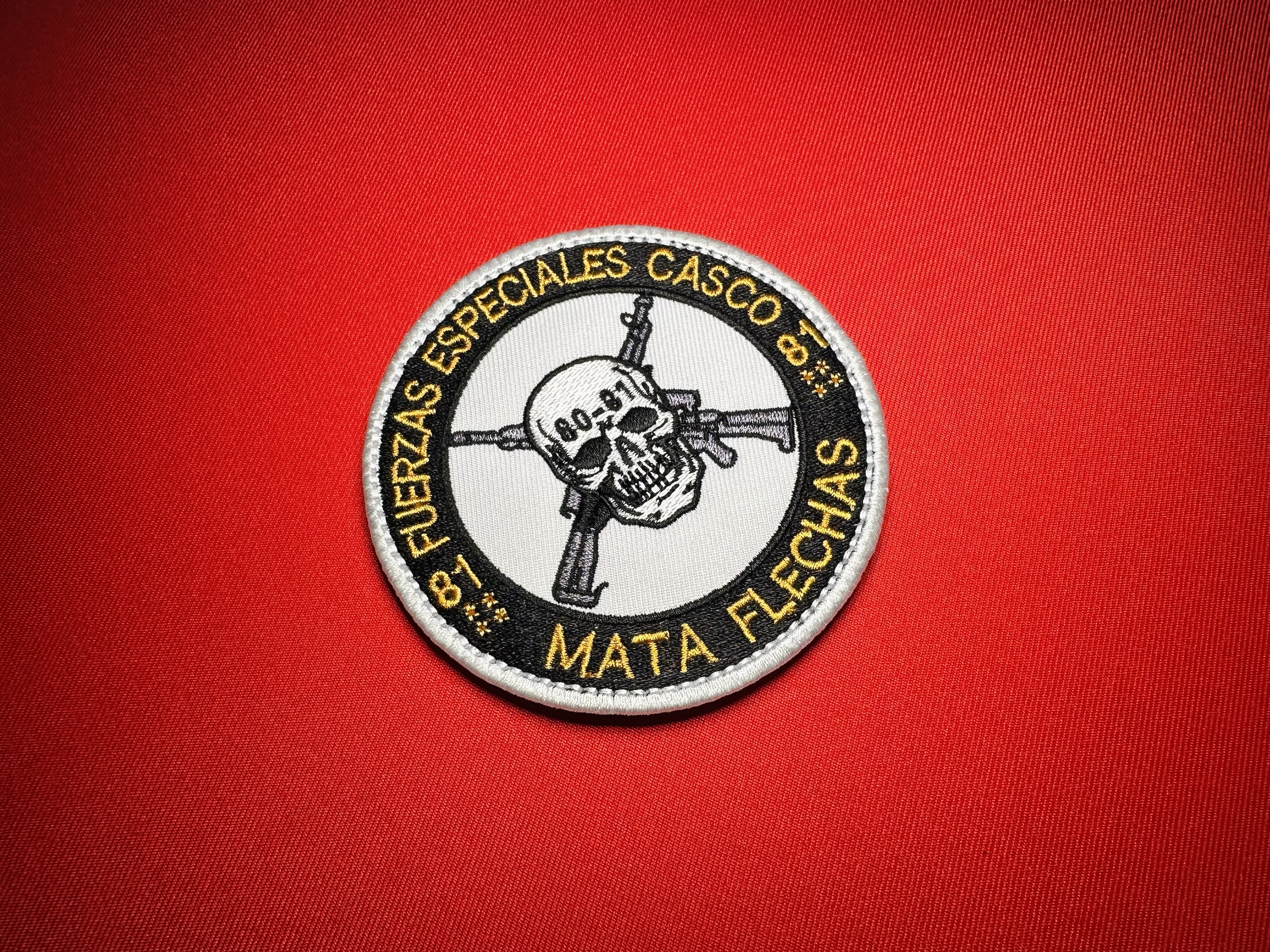 Angled view of the Mata Flechas Fuerzas Especiales Casco reproduction patch, showing gold lettering, a skull marked 80-81, crossed rifles, and embroidered details.