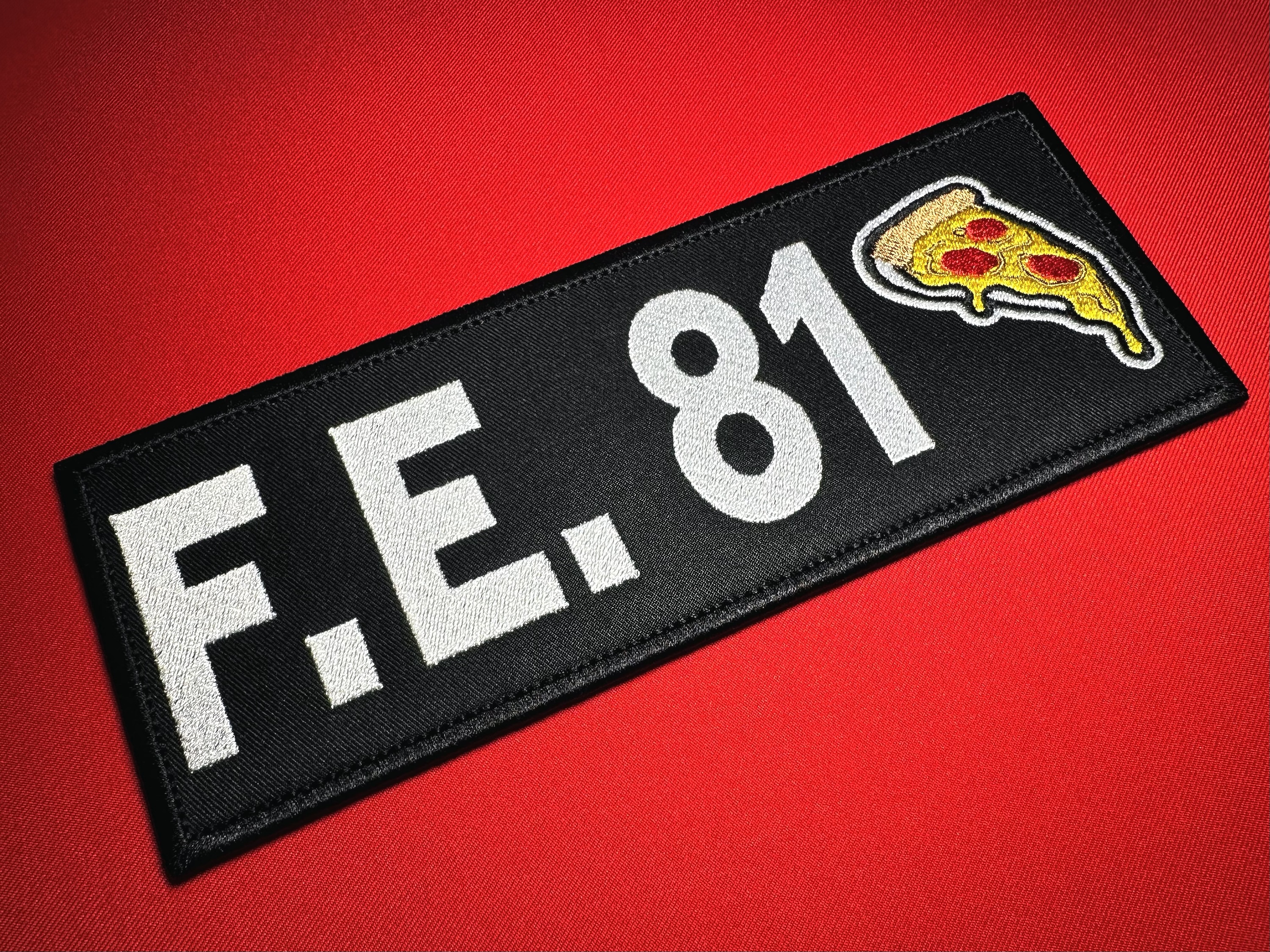 Angled view of a 9 × 3.5-inch F.E. 81 La Chapiza reproduction patch with white embroidered lettering and a pepperoni pizza emblem on black, displayed on red fabric.