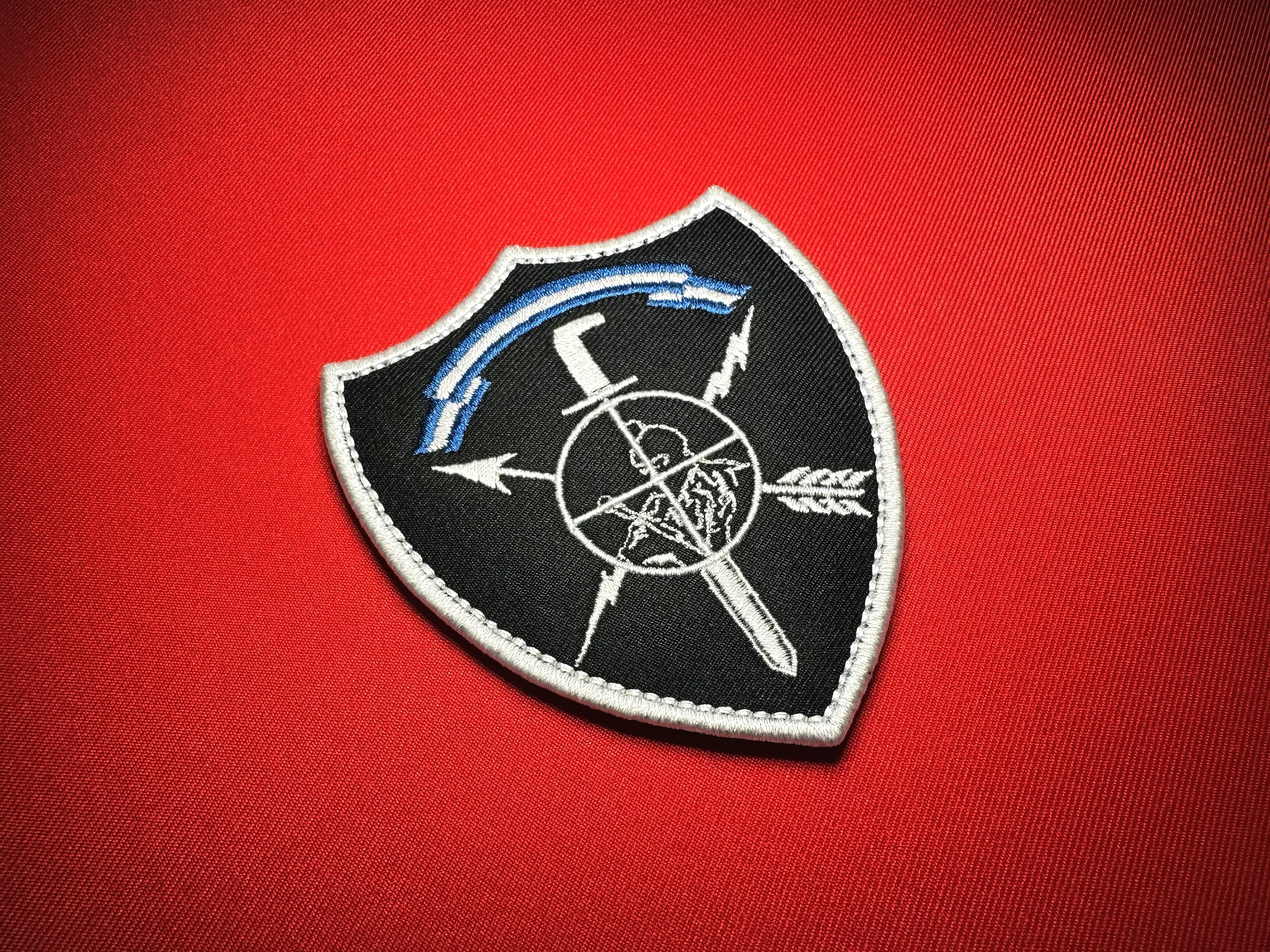 Angled close-up of the El Salvador Comando Especial Antiterrorista (CEAT) replica patch, showing detailed white embroidery, a black shield and white border on red fabric.