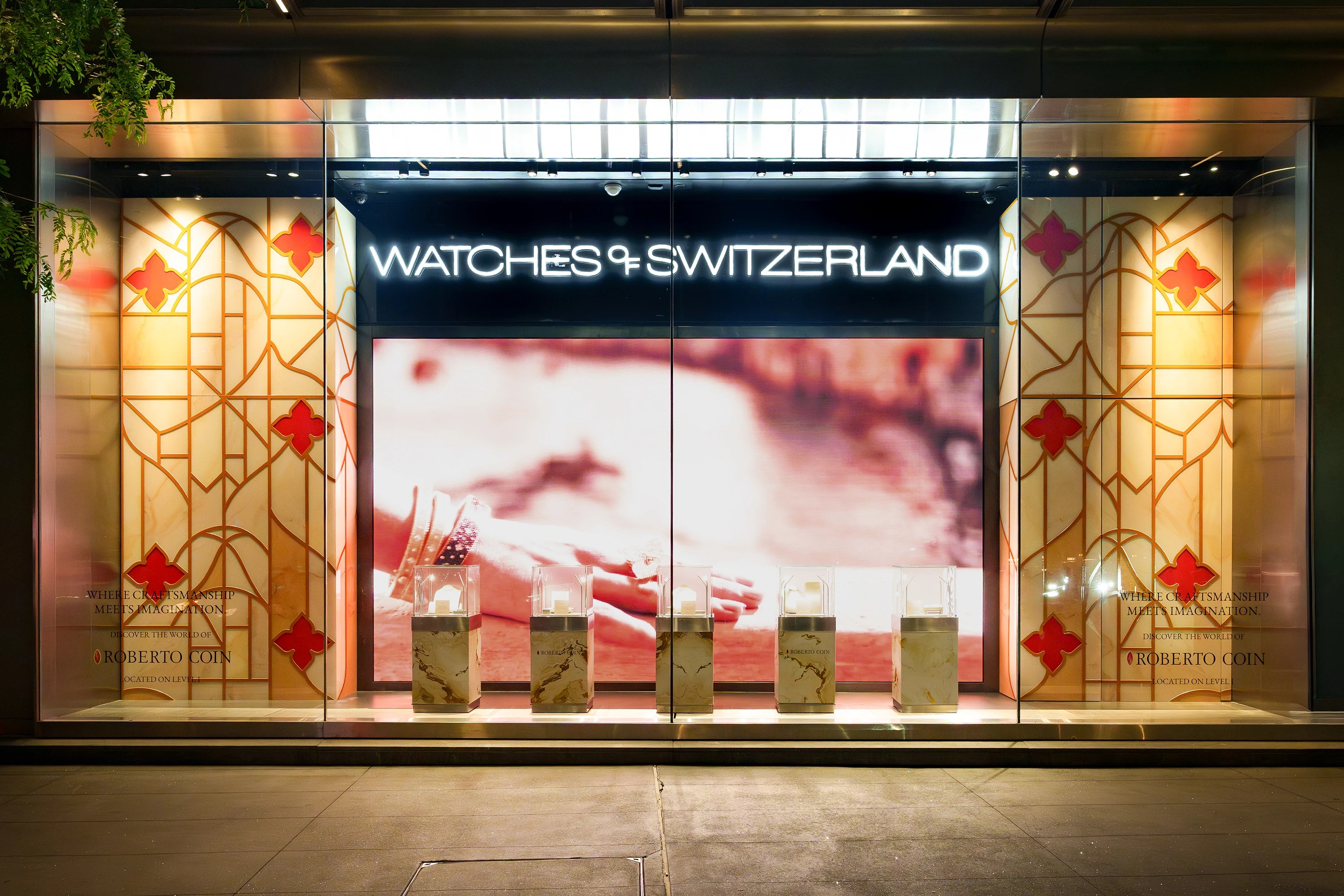 Watches of Switzerland Hudson Yards window display with illuminated neon fascia, Roberto Coin large-format backlit graphic, and luxury marble display pedestals