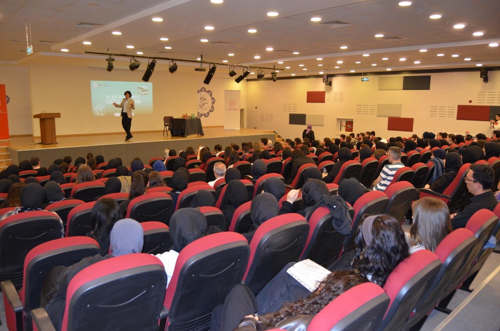 Bir sunum yapılan büyük bir konferans salonunda, ön planda bir konuşmacı sahnede duruyor. Arkada birçok izleyici, sandalyelerde oturmuş dikkatlice dinliyor.
