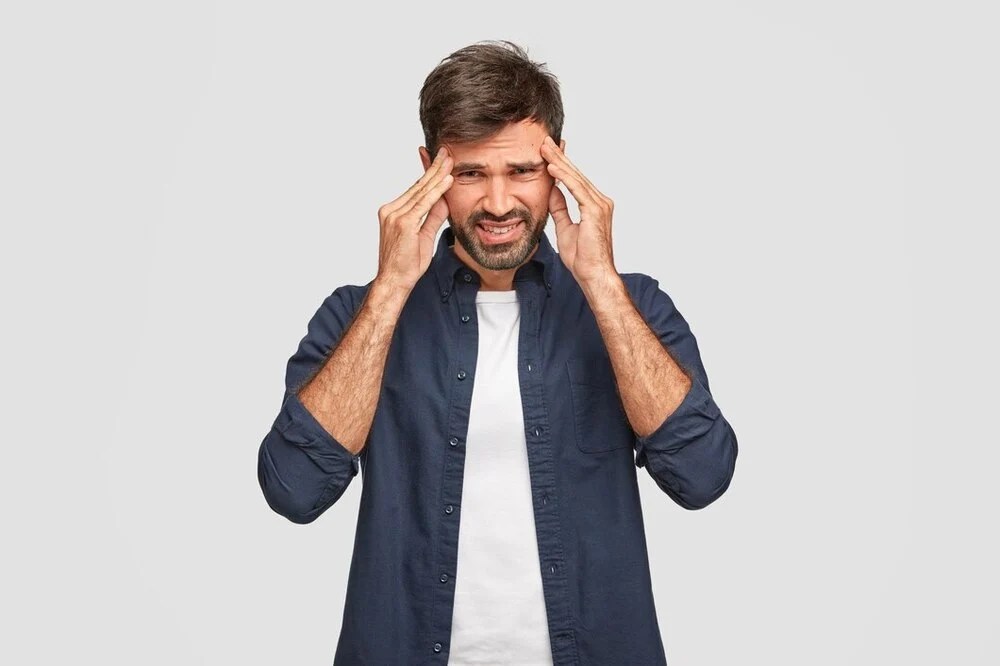 Person Holding Their Temples While Standing Against A Plain Background