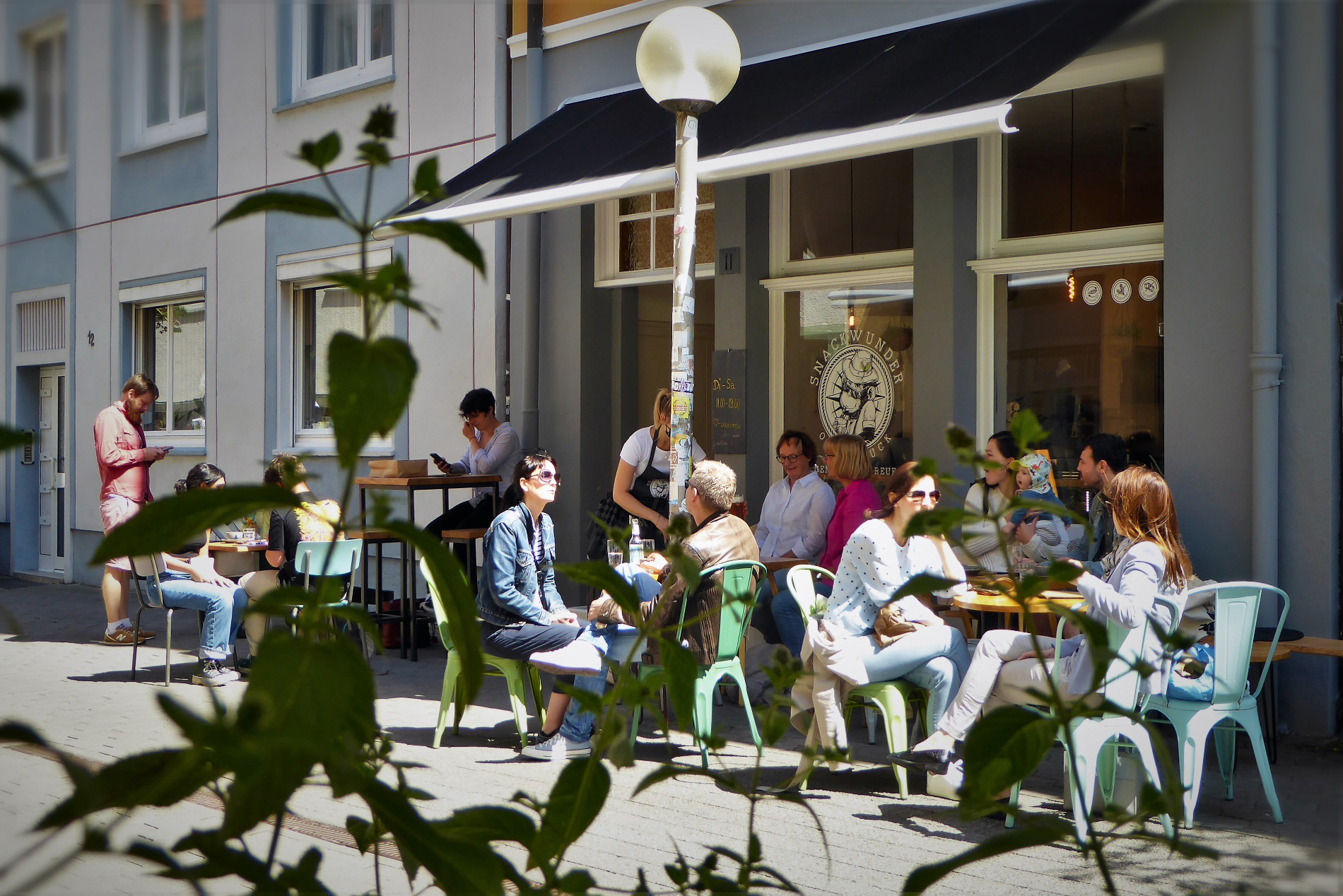 Blick auf die Außentische des Snackwunders: Menschen sitzen in der Sonne auf bunten Metallstühlen an kleinen Tischen unter der Markiese. Die Sonne scheint.
