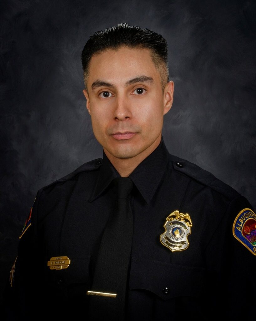 A uniformed law enforcement officer stands in formal attire with a badge and patches visible against a dark background.