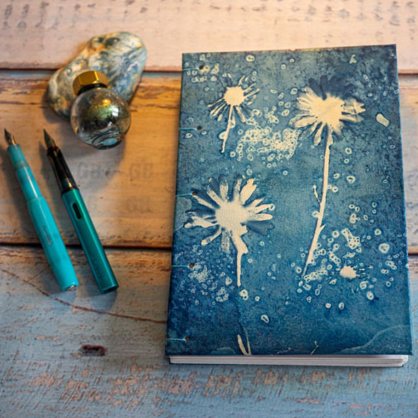 A hand bound journal with a cyanotype print cover, with daisies. There are fountain pens and an ink bottle