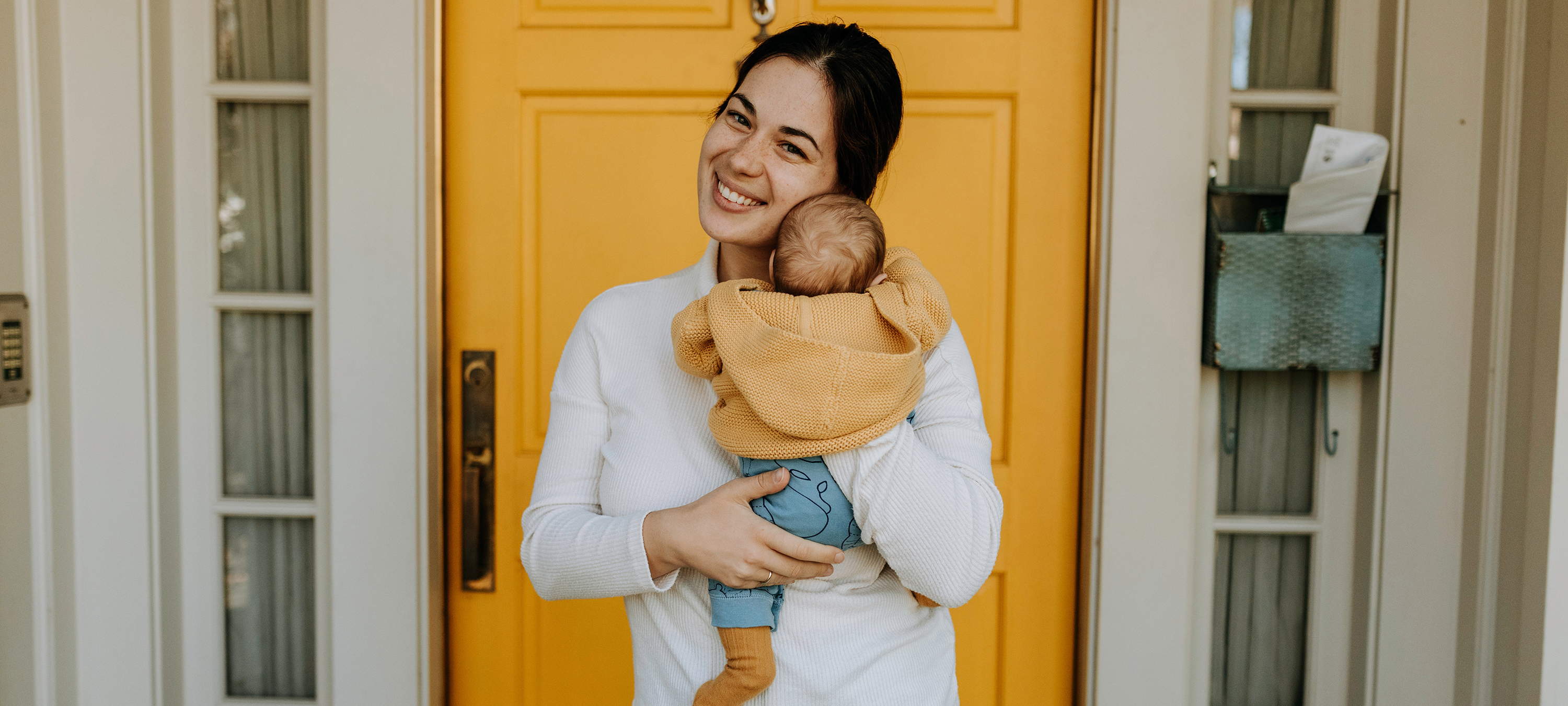 woman smiling, holding a baby, in front of a yellow door
