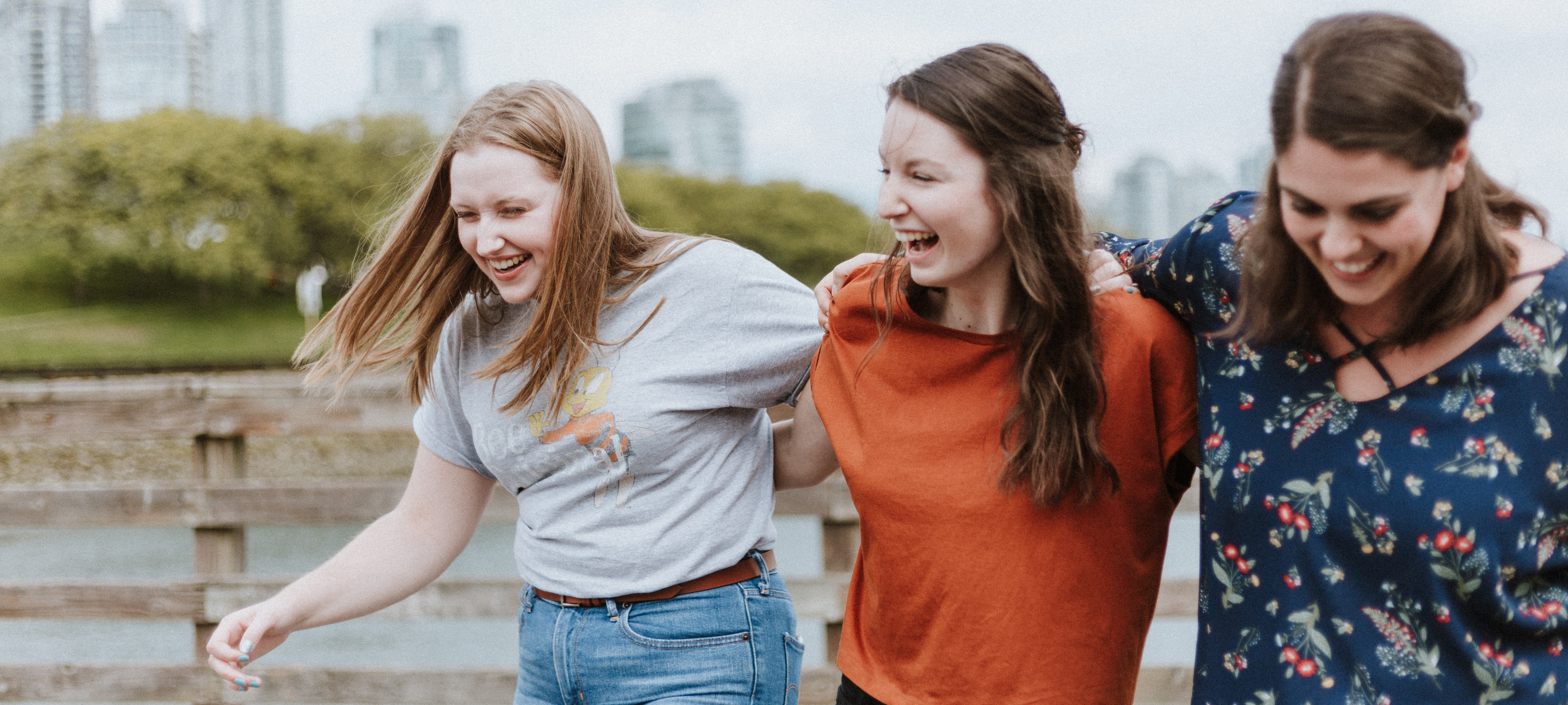 three female friends, smiling with arms wrapped around each other near a river