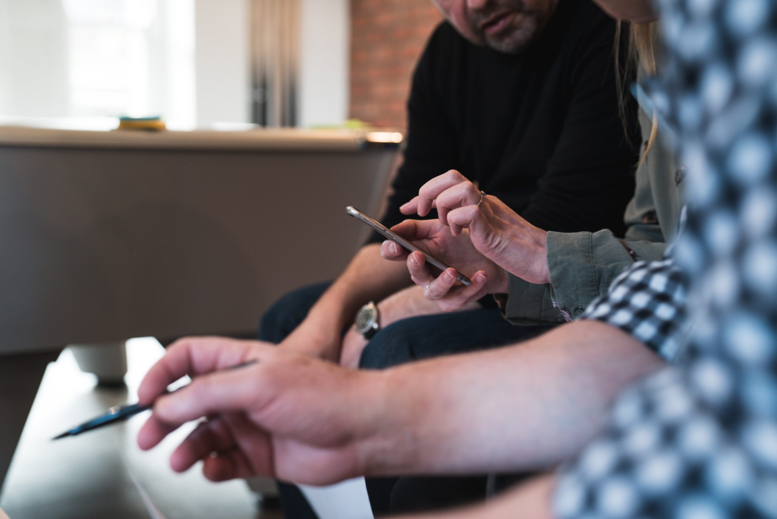 Drei Menschen sitzen nebeneinander, die Person in der Mitte hält ein Smartphone in der Hand, eine andere schaut ihr auf den Bildschirm, die dritte Person hält einen Stift in der Hand.