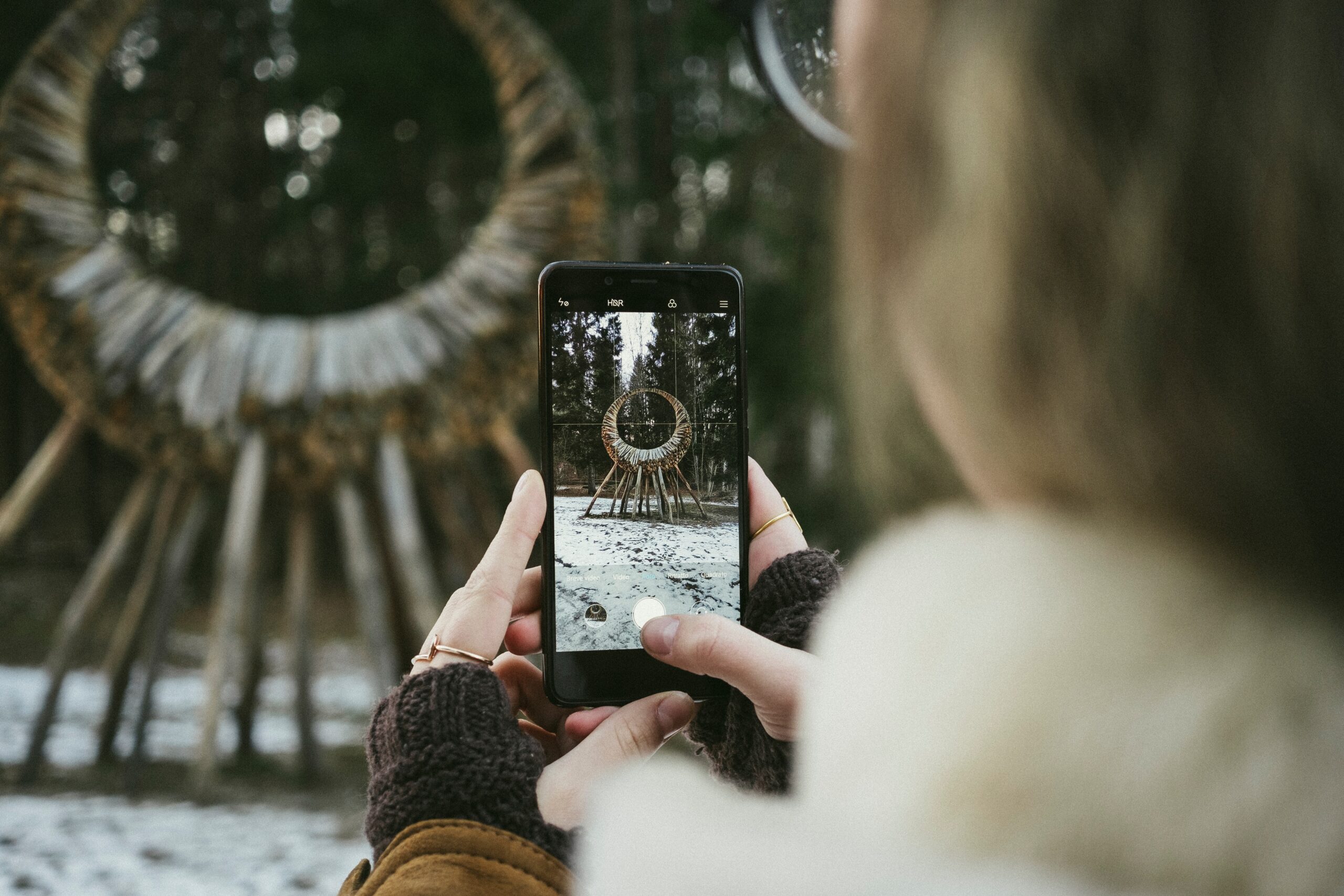 Frau hält Smartphone in den Händen und nimmt ein Foto auf von einem Bauwerk aus Holz am Waldrand.