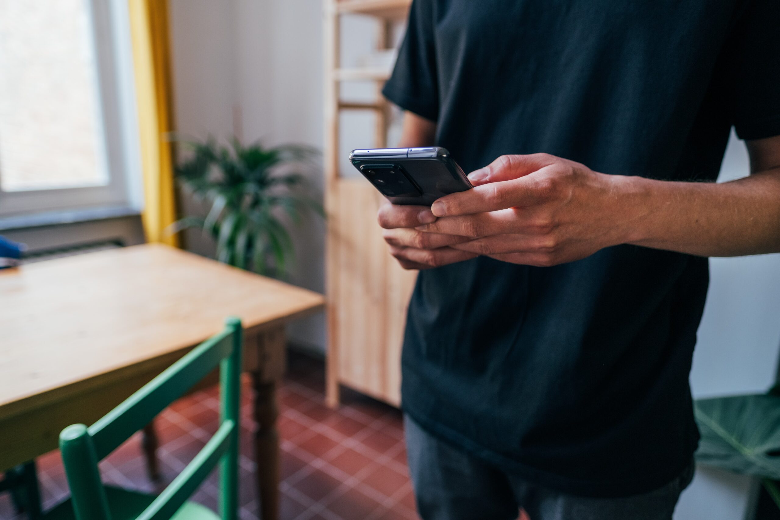 Mann in Wohnzimmer mit Smartphone in den Händen. Die Daumen sind auf dem Bildschirm.