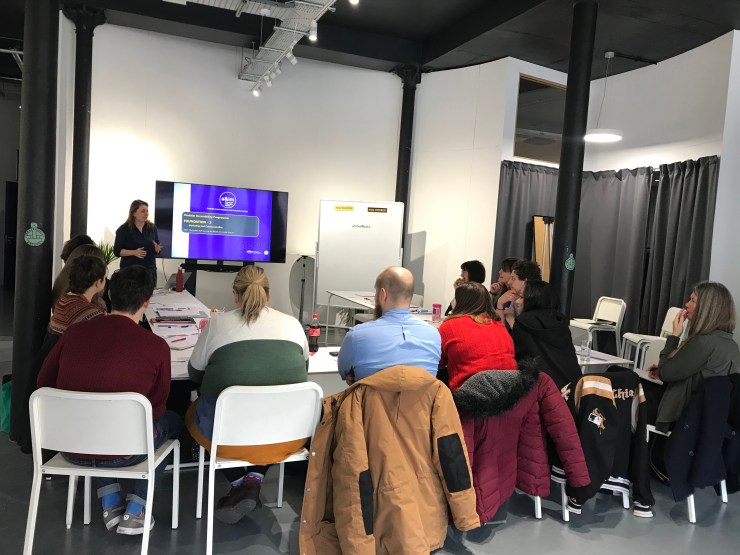 the photo shows a training room set up with participants seated around tables. Trish is standing at the front alongside a large screen TV which has the introduction slide from the training presentation on it