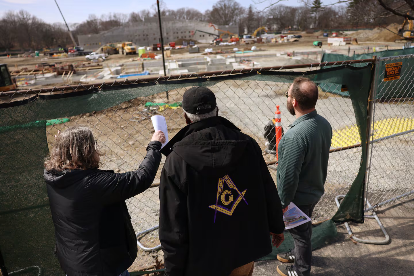 Three people are seen from behind, looking through a chain-link fence at construction site