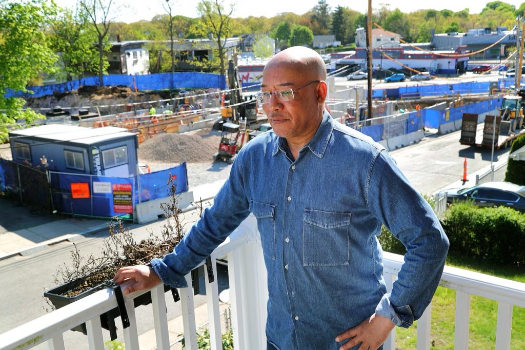 A man with bald head, rimless glasses, dark skin and a blue denim shirt stands in the sun in front of a construction project