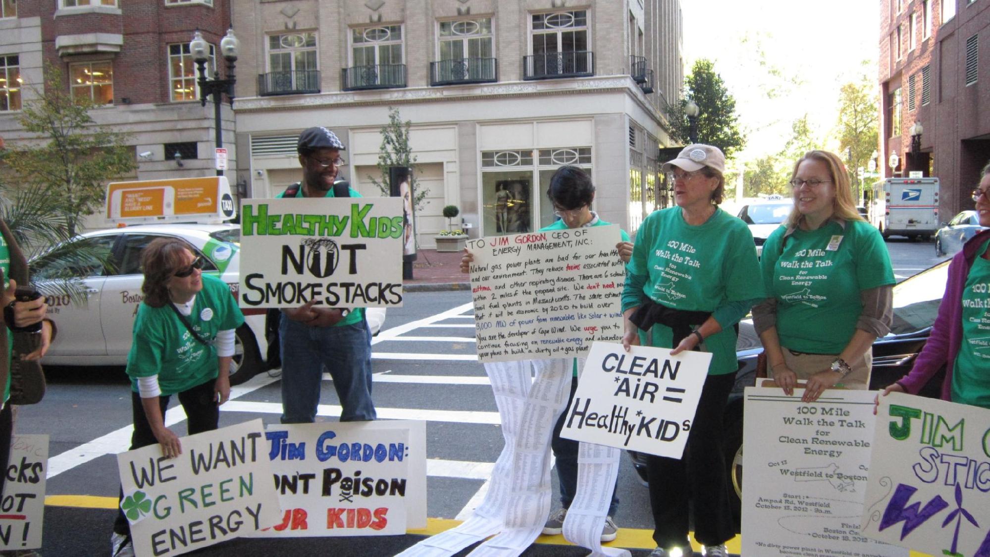 a group of people in green t-shirts stand outside on a Boston street holding protest signs