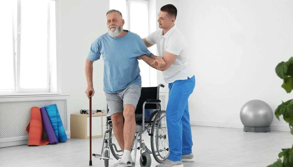 Person being assisted by a healthcare professional to stand from a wheelchair in a neurological rehab room