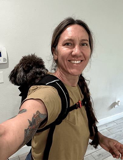 Woman smiling in a selfie with a small dog carried in a backpack.