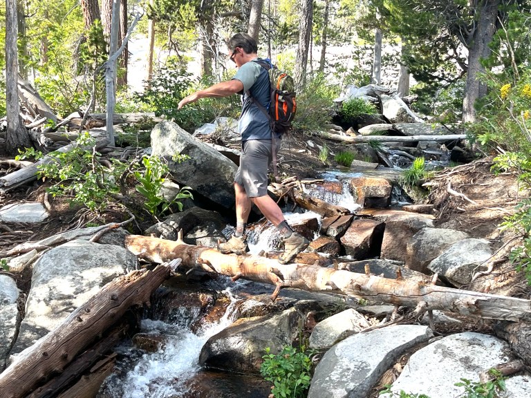 A hiker stepping across fallen logs over a rocky creek in the Sierra backcountry