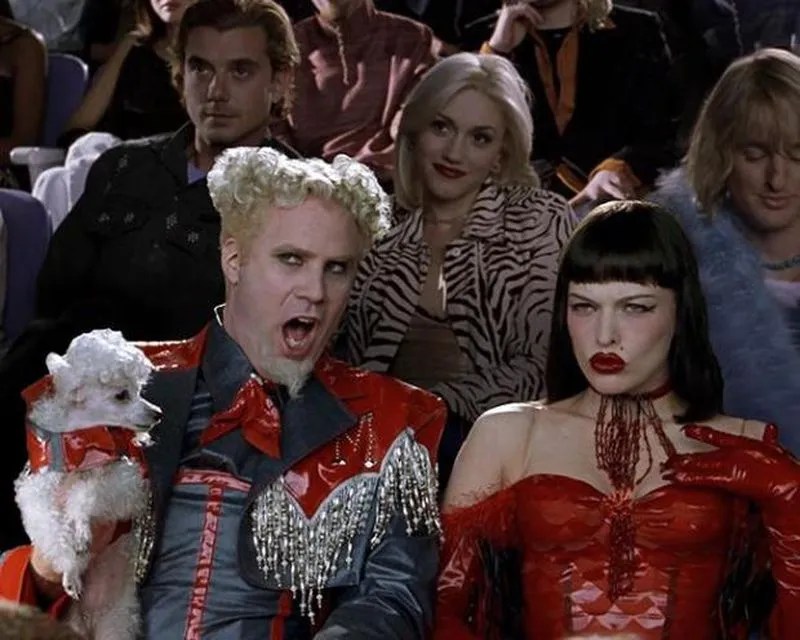 A scene from a movie featuring two fashionable characters with distinct hairstyles and outfits, sitting in a crowd, with one holding a small dog.