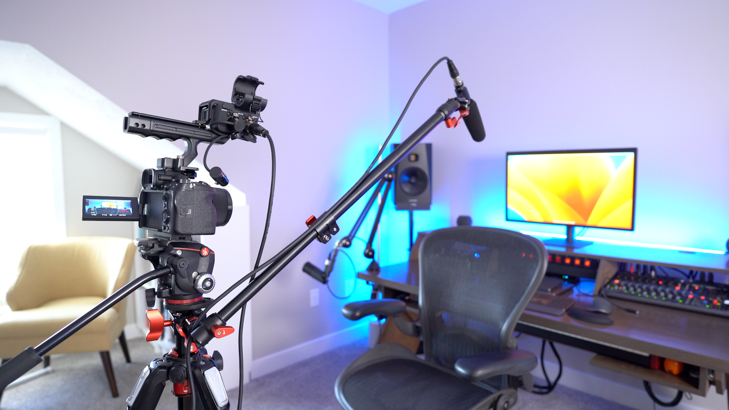 A professional video or streaming setup with a camera on a tripod, boom arm with microphone, computer monitor, sound mixing console, and a desk chair in a studio or home office with blue accent lighting.