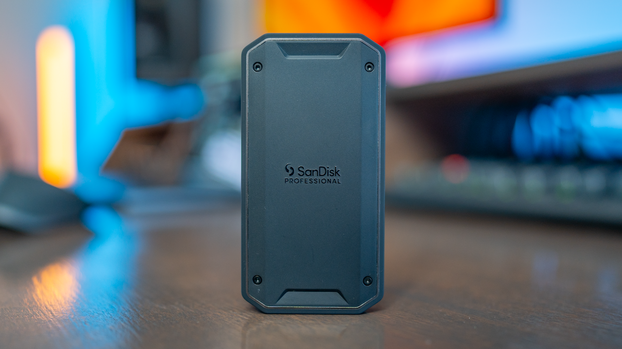 A black SanDisk Professional external hard drive standing vertically on a wooden desk, with colorful blurred computer monitors in the background.