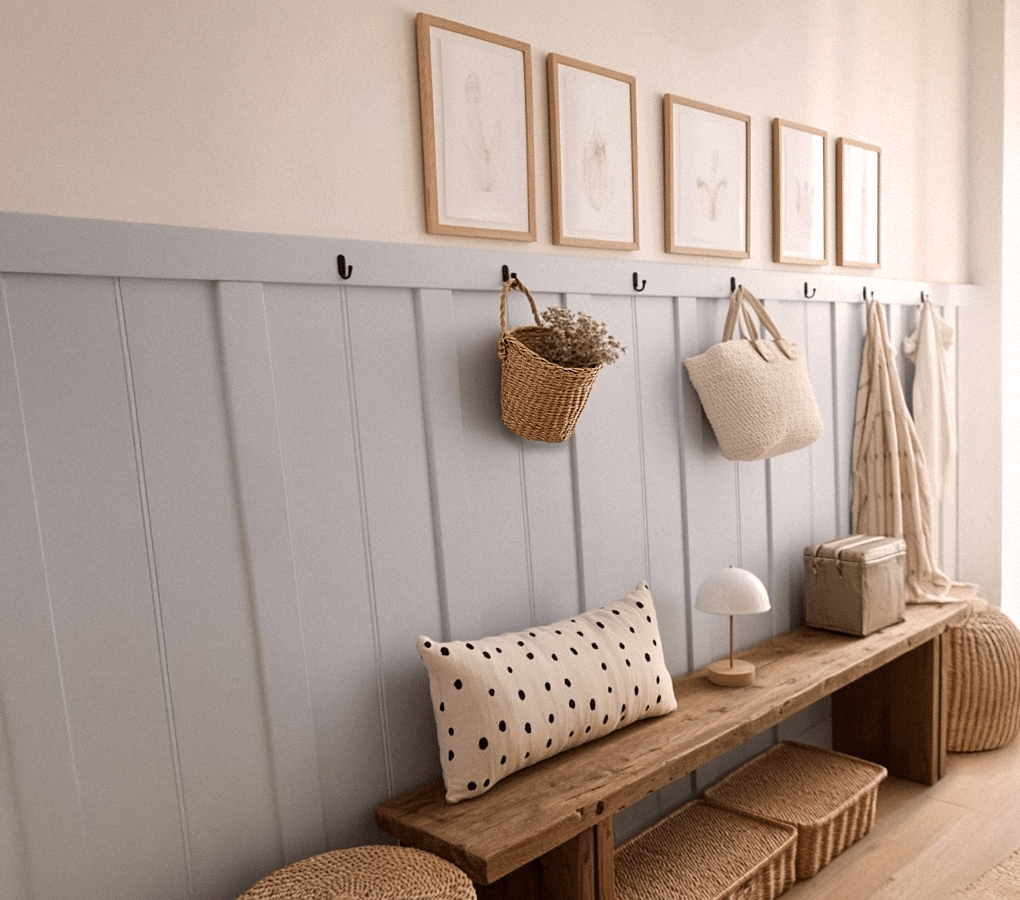 Banc en bois naturel sous un mur bleu-gris avec patères et paniers suspendus dans une petite entrée lumineuse et chaleureuse.