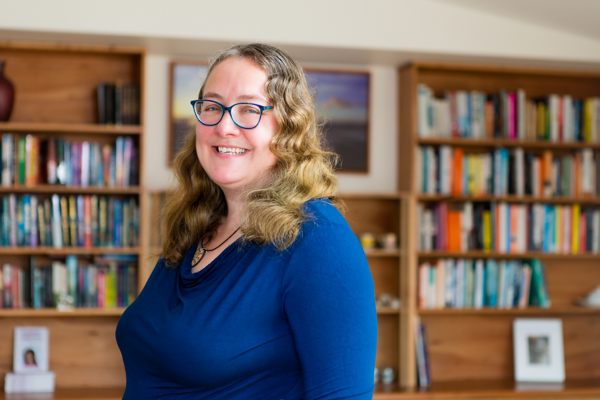 Photo of Linda McIver, in front of a bookshelf, smiling at the camera, in a dark blue long sleeved top