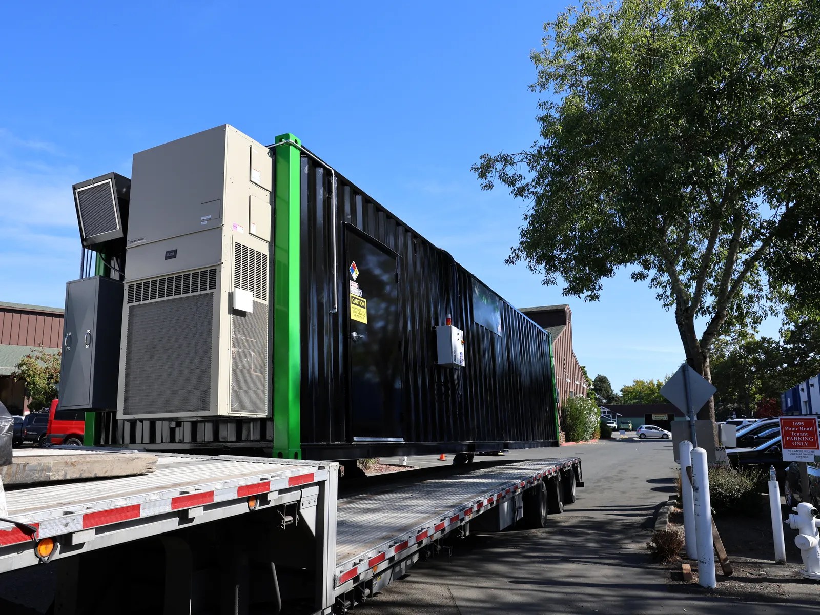 Black modular container with green trim and end-mounted HVAC loaded on a flatbed trailer