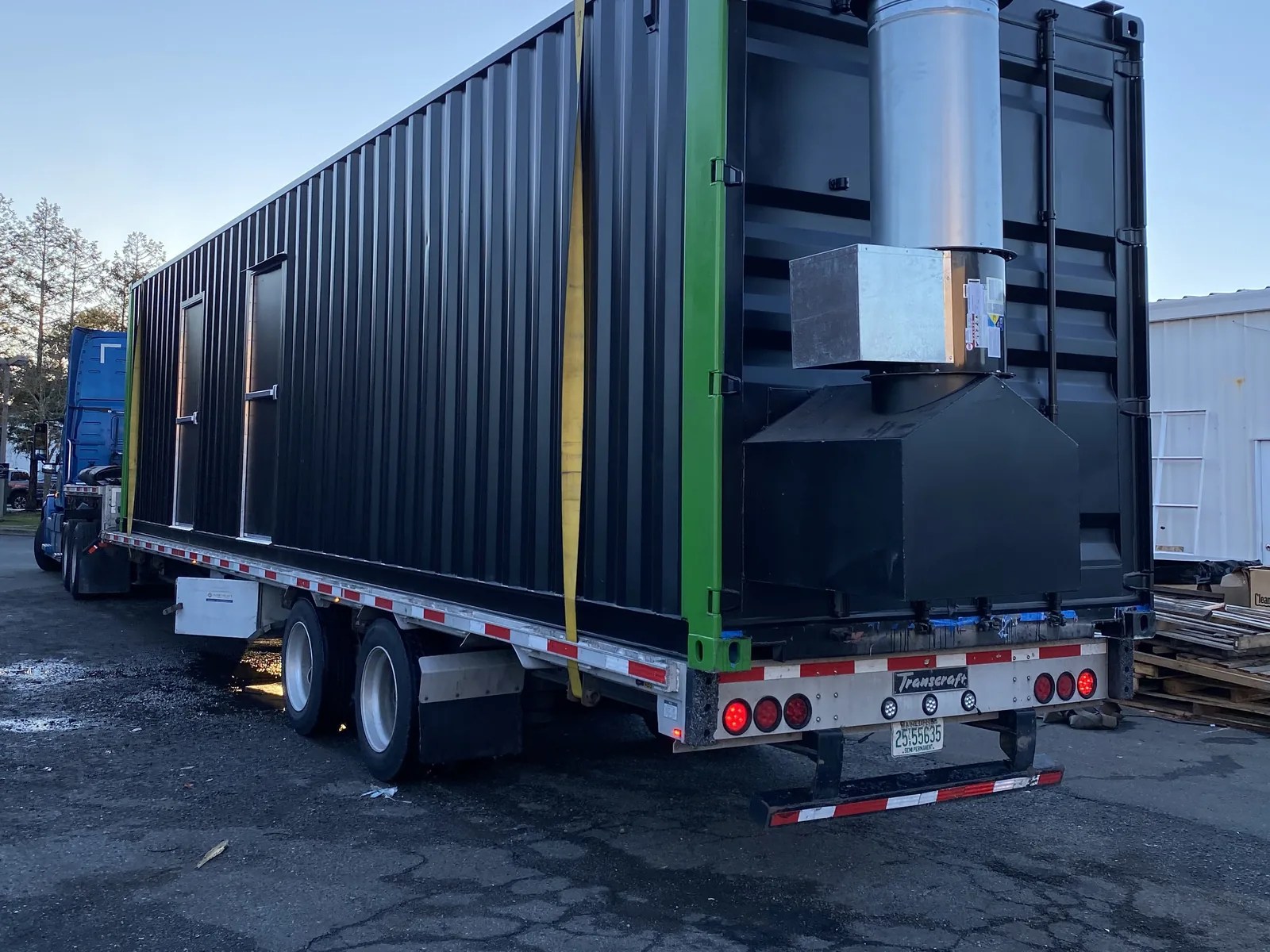 Black 40 ft container room with green corner posts and an exhaust stack on a flatbed trailer, rear view