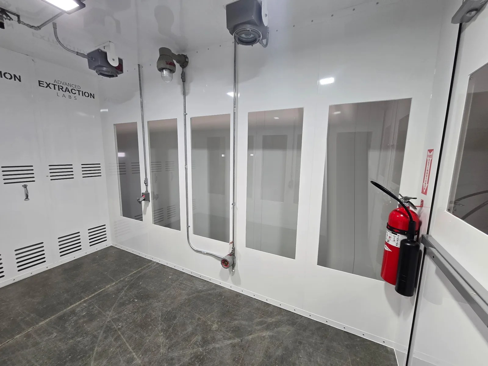Walk-in booth interior with white wall panels, vision windows, louvered vents, conduit and a fire extinguisher