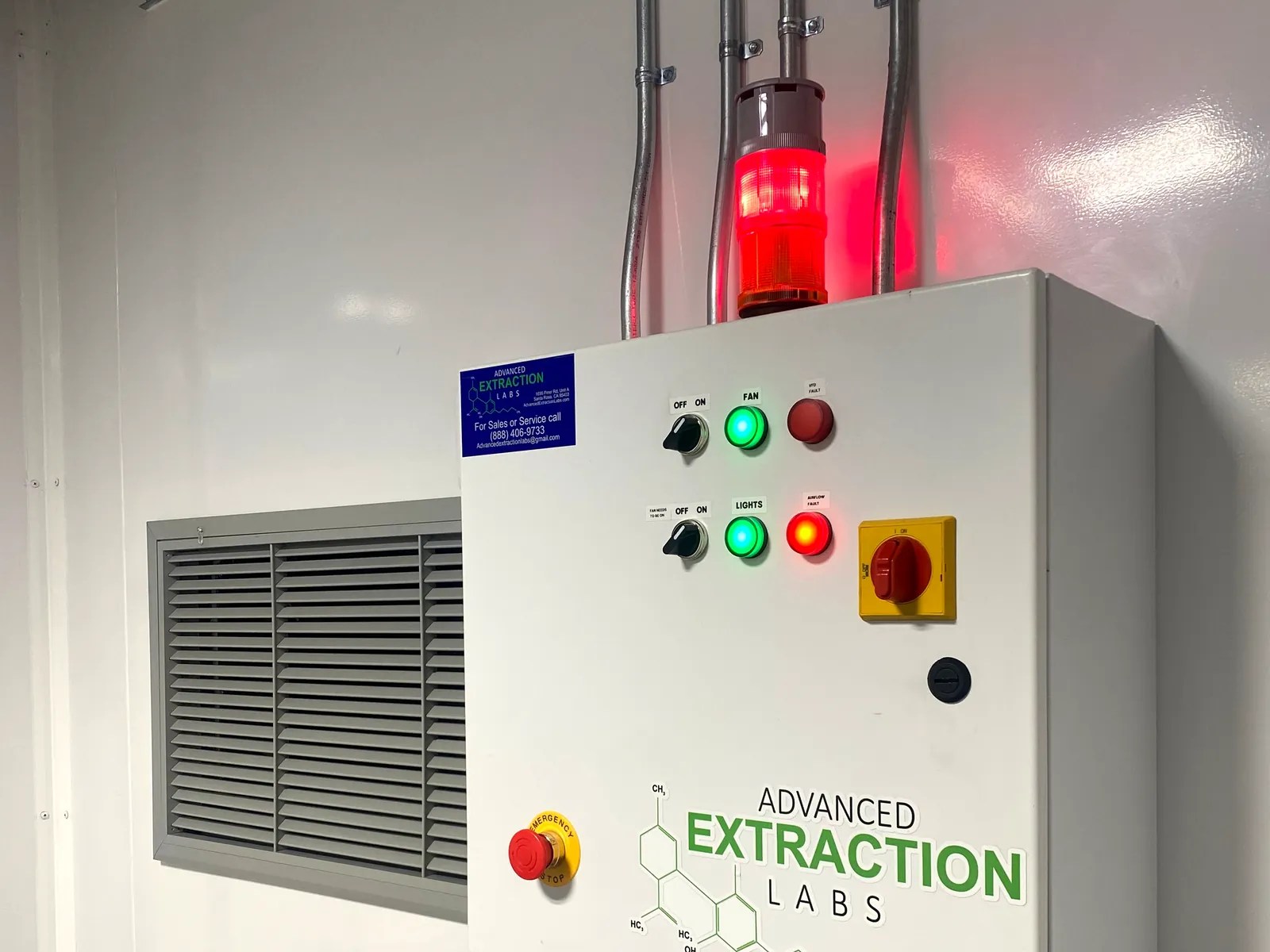 Wall-mounted control panel with a red beacon and indicator lights beside a louvered vent inside a white room