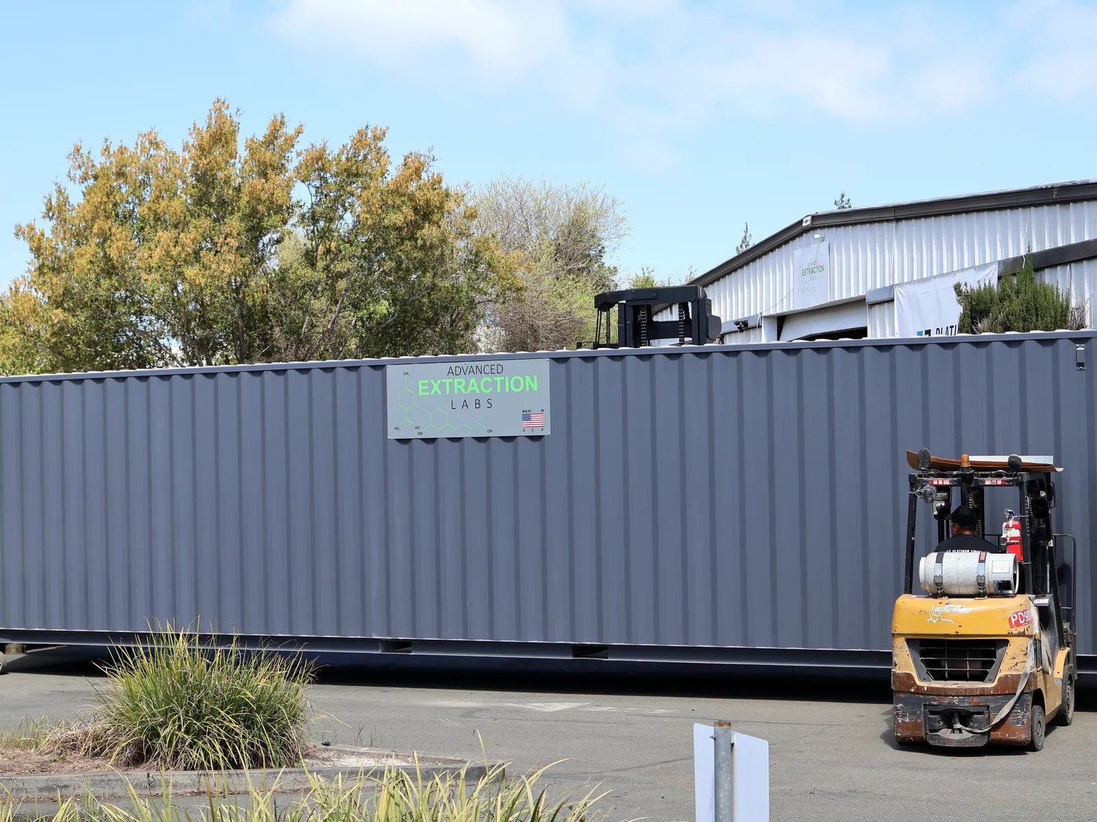 Long grey modular container with green trim being moved by a forklift in a sunny yard