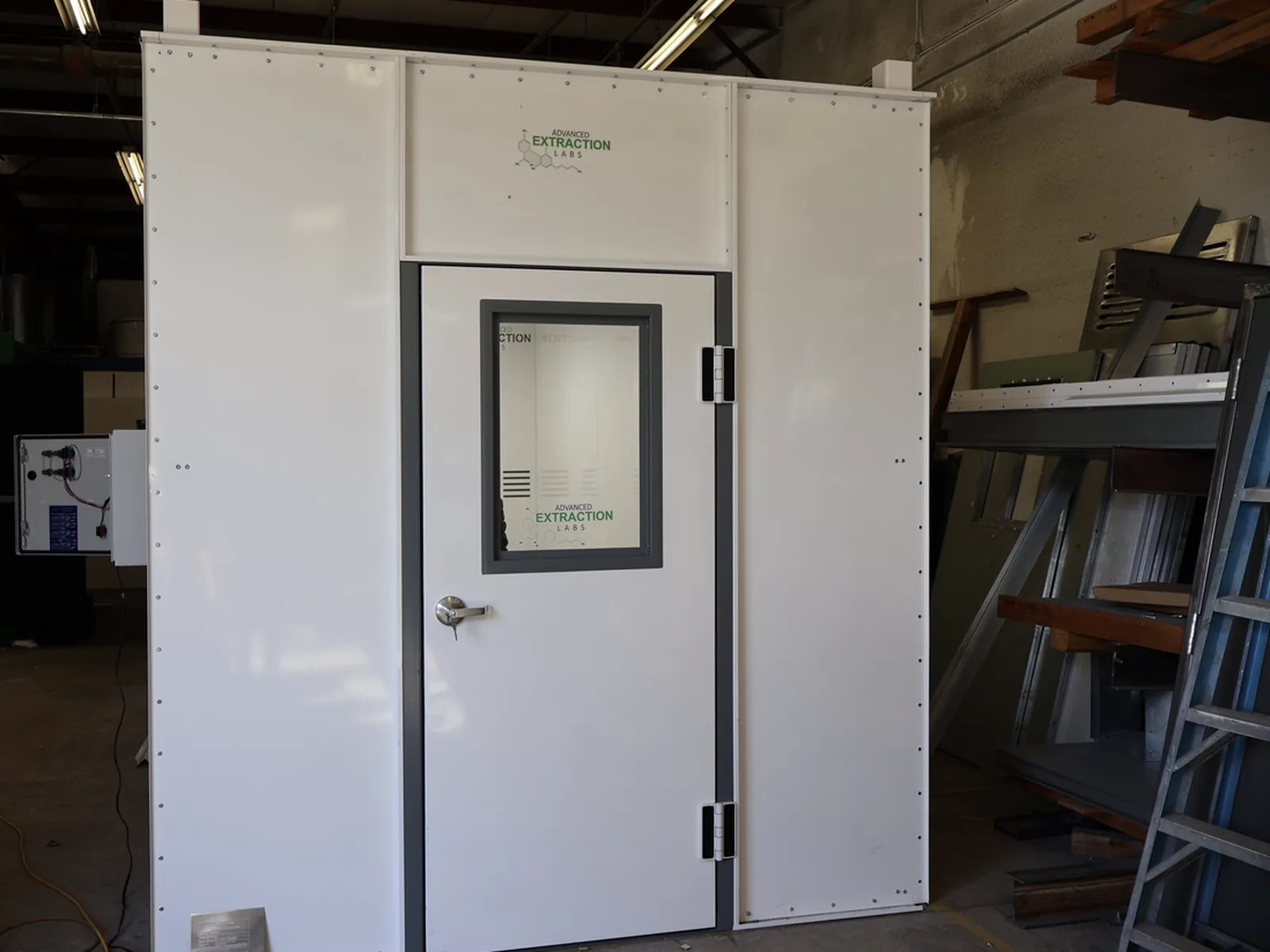 White single-door walk-in booth with a vision window and an exterior control box in a fabrication shop