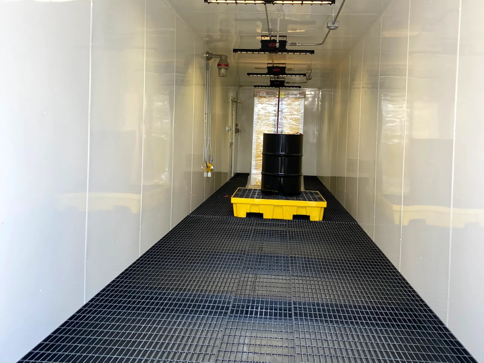 White storage container interior with steel floor grating over a containment sump, lighting and a spill pallet at the far end